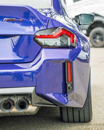 Close-up rear three-quarter view of a glossy blue BMW M2, showing the taillight, rear diffuser, exhaust pipes, and a blurred vehicle in the background.